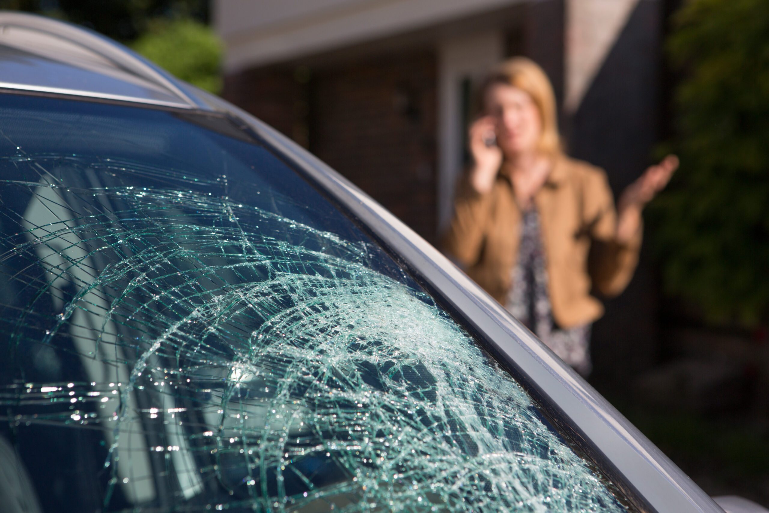 A car with a shattered windshield in the foreground with a woman talking on the phone in the background.